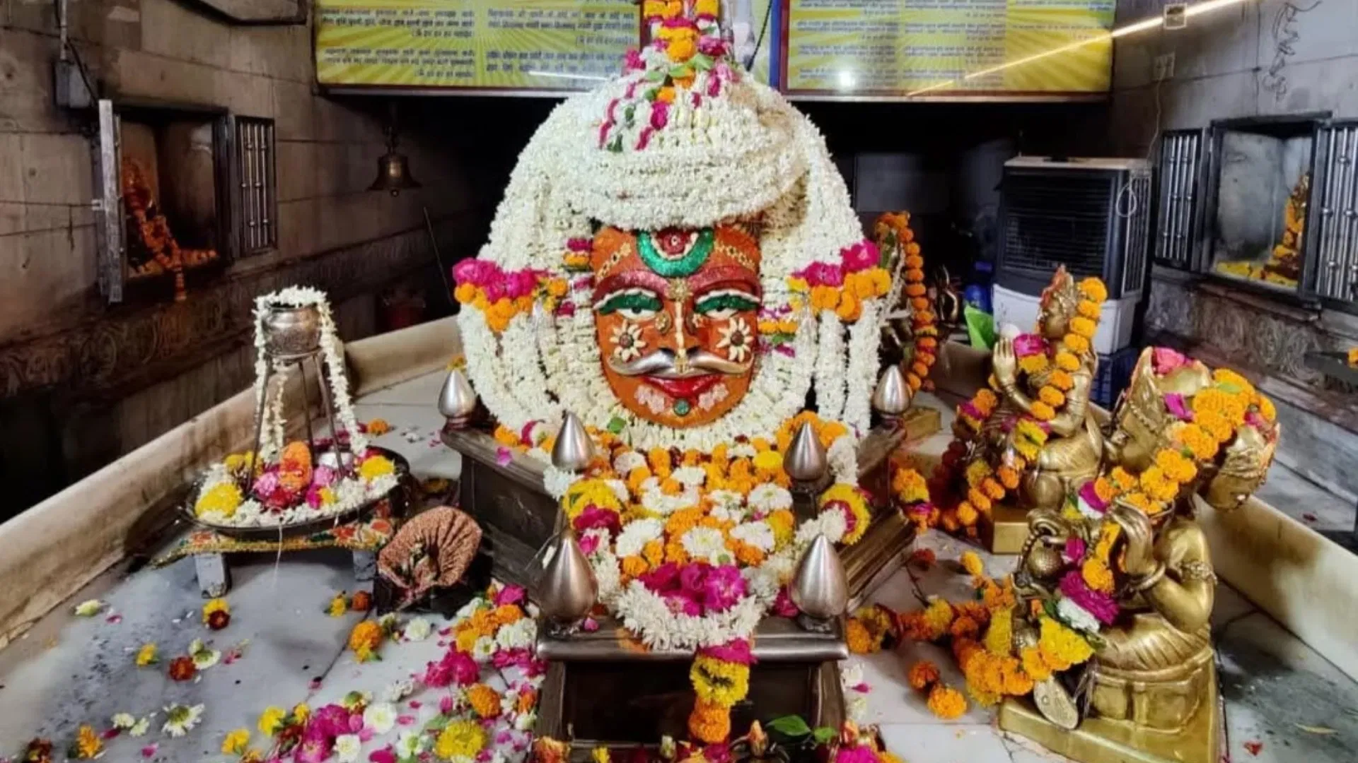 The linga in full shringar — its painted silver face heaped with jasmine and marigold garlands, brass idols to one side